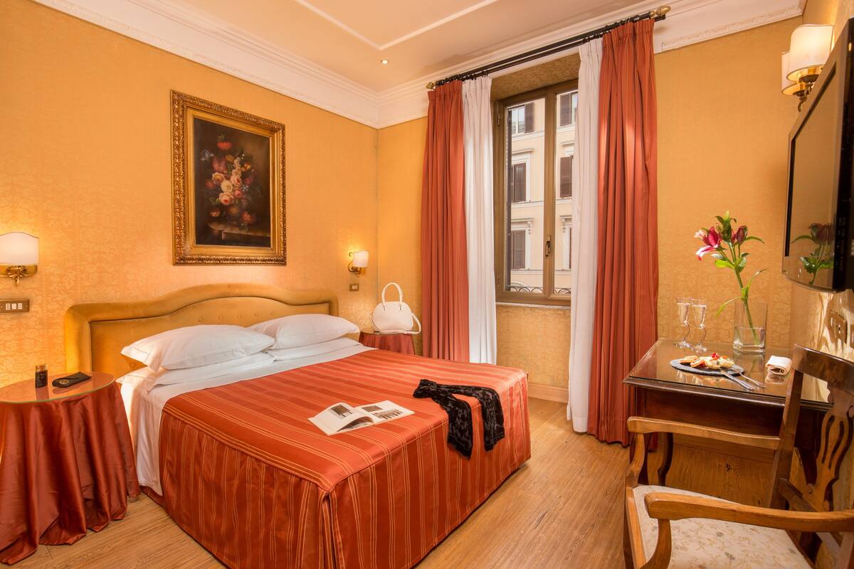 A hotel room with a large bed, a desk with a chair, a window with curtains, a painting on the wall, and a lamp on the nightstand.