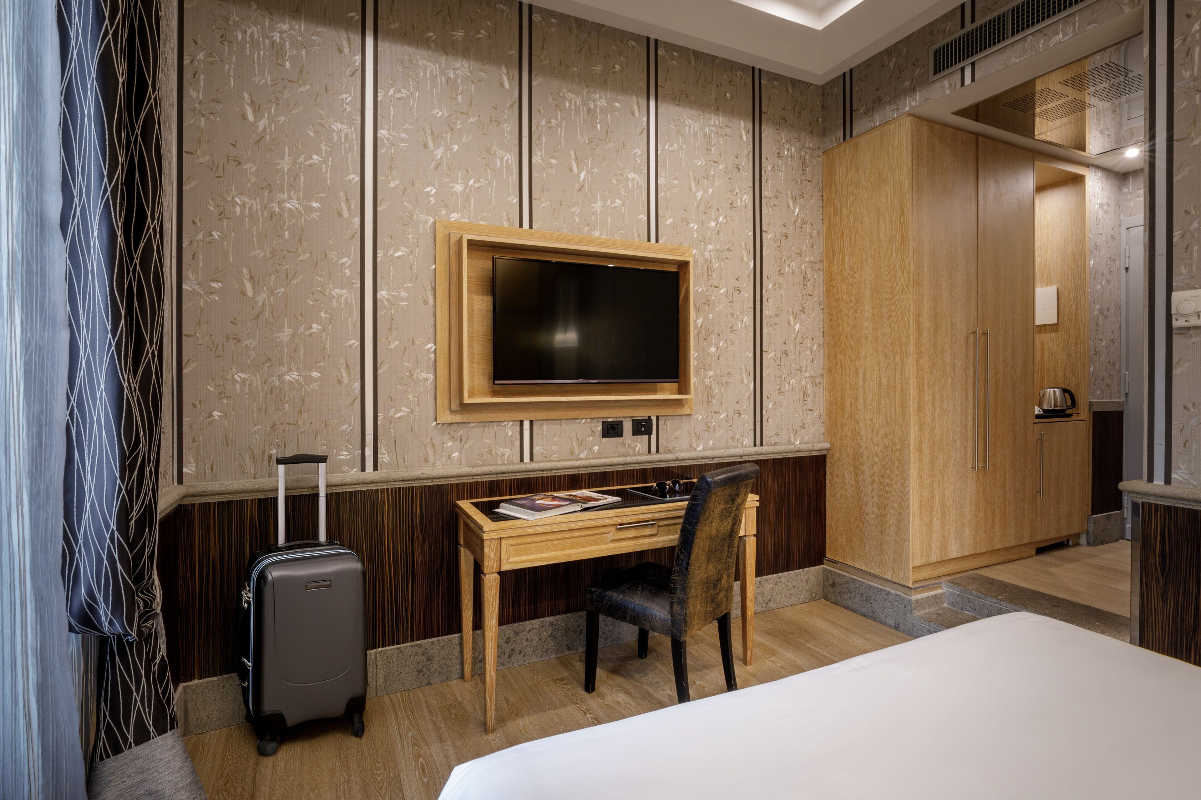 A hotel room with a bed, a desk, a chair, a TV mounted on the wall, and a suitcase.