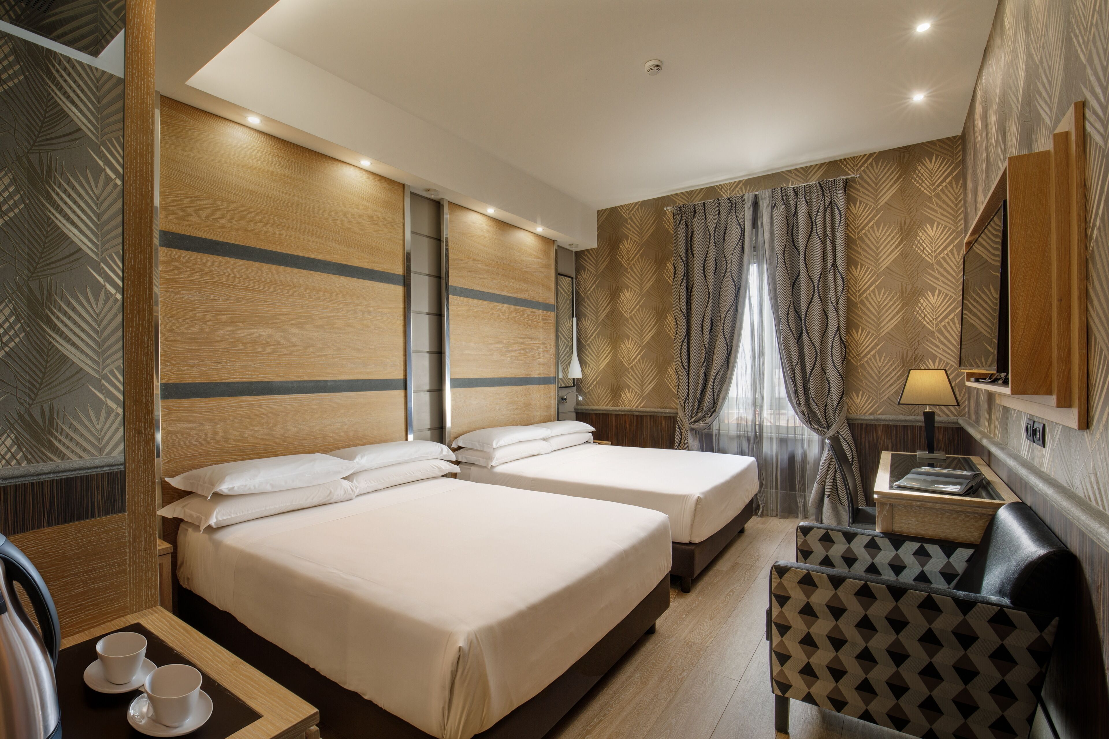 A hotel room with two beds, a small table with a cup and saucer, a chair, and a nightstand with a lamp.