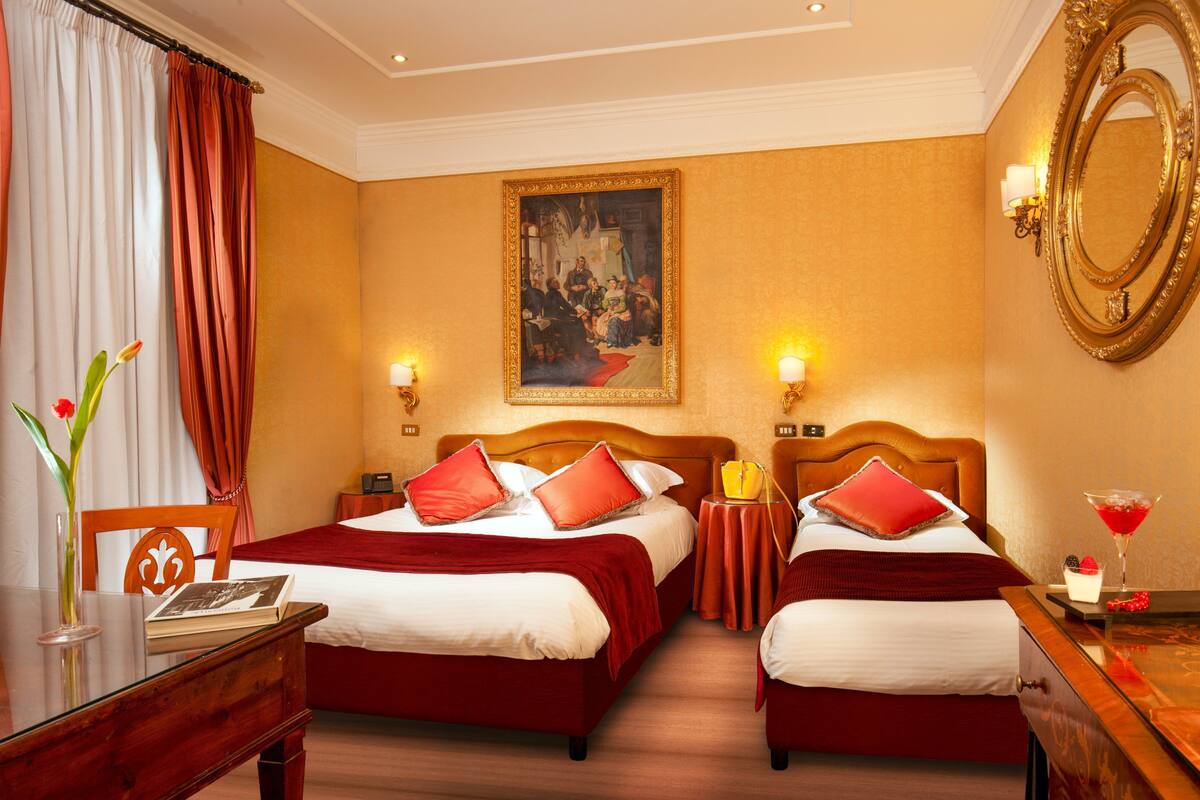A hotel room with two beds, a painting on the wall, a mirror, and a desk with a vase of flowers.