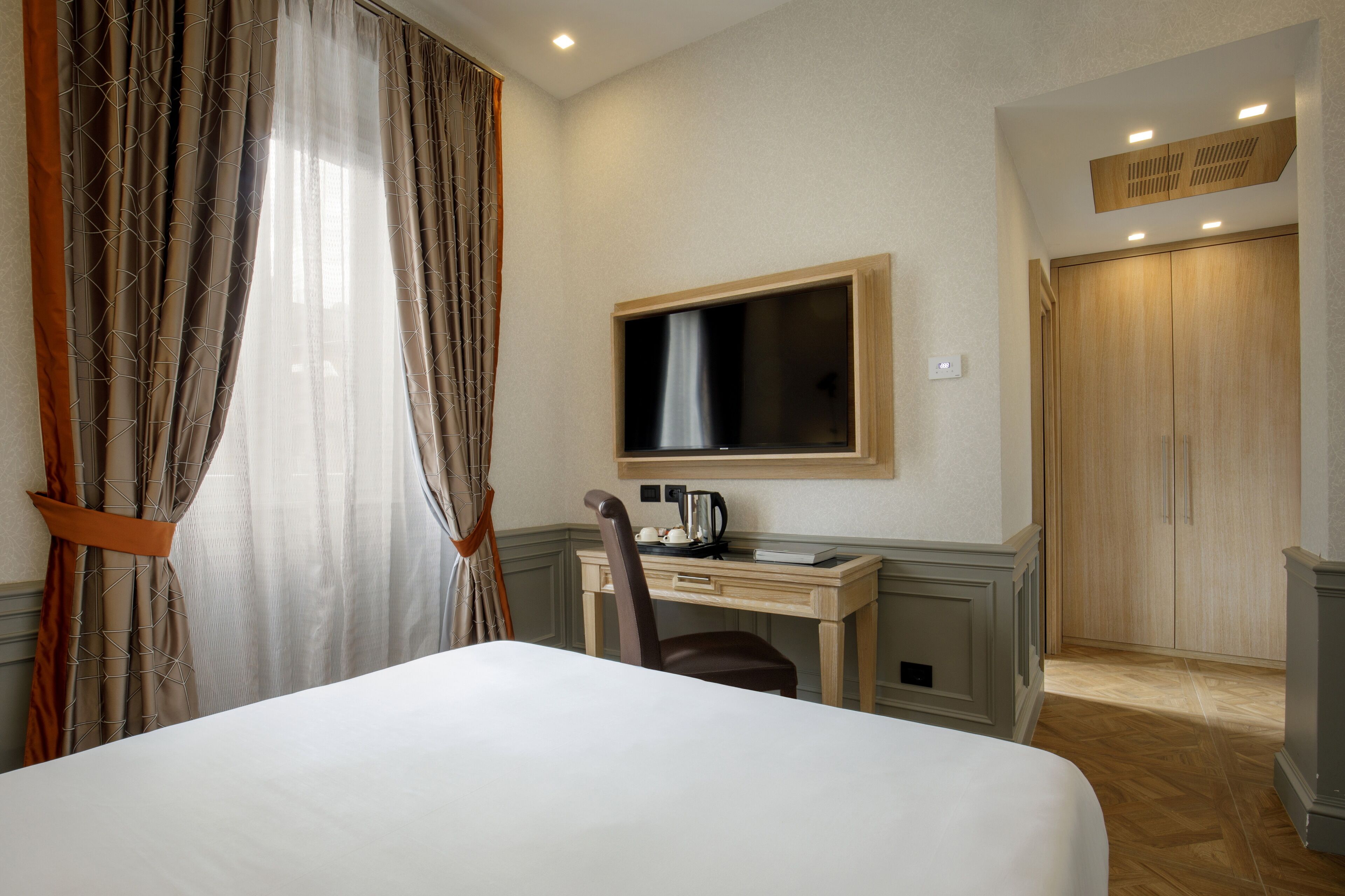 Habitación de hotel con cama, escritorio con silla, televisor, armario y ventana con cortinas.
