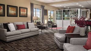A spacious living area with a sofa set, armchairs, and a coffee table. There are framed pictures on the wall, a large window with curtains, and a bookshelf filled with books and decorative items.