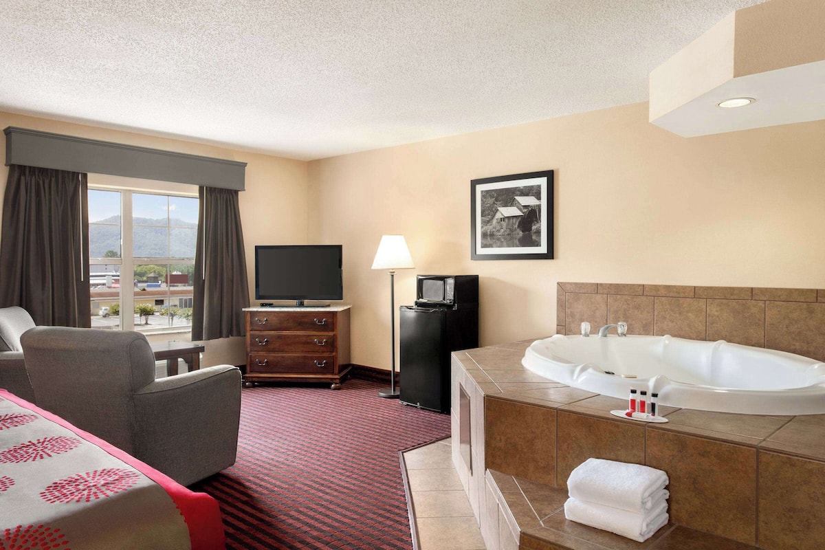 Habitación de hotel con una cama grande, un sofá, un televisor, un pequeño televisor, una bañera y una ventana con cortinas.