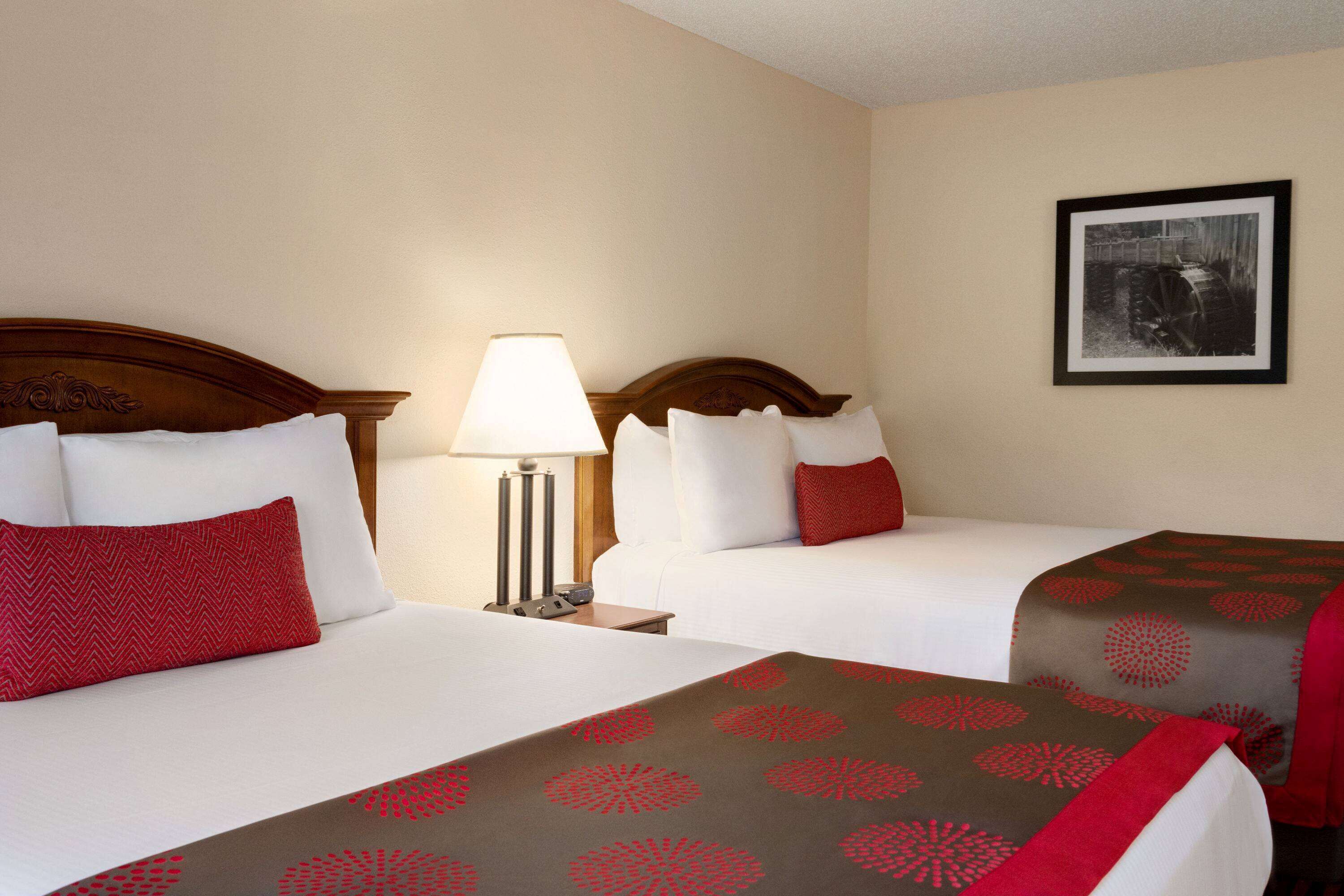Habitación de hotel con dos camas, cada una con almohada roja y blanca, una mesita de noche con lámpara y un cuadro en la pared.