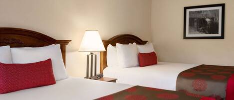 Habitación de hotel con dos camas, cada una con almohada roja y blanca, una mesita de noche con lámpara y un cuadro en la pared.