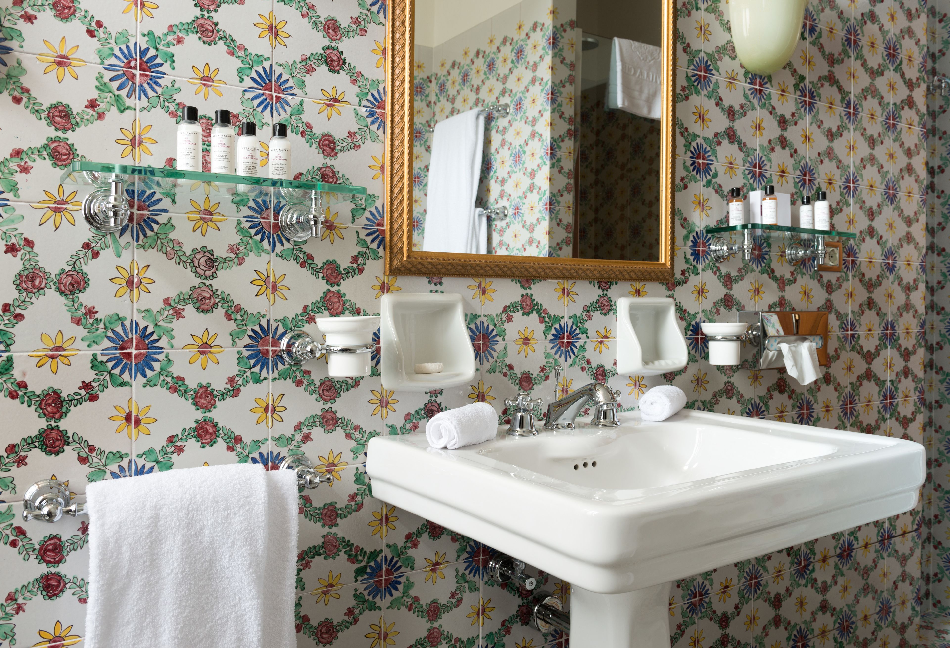 A bathroom with a floral wallpaper, a white sink, a mirror, and shelves with toiletries.