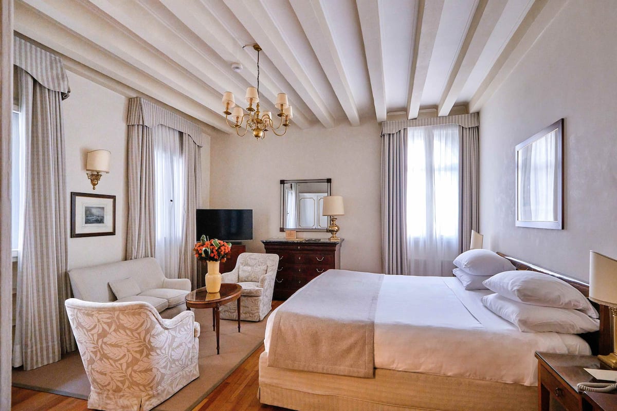 A hotel room with a large bed, a sofa, a chair, a small table with a vase, a dresser, a mirror, and a television.