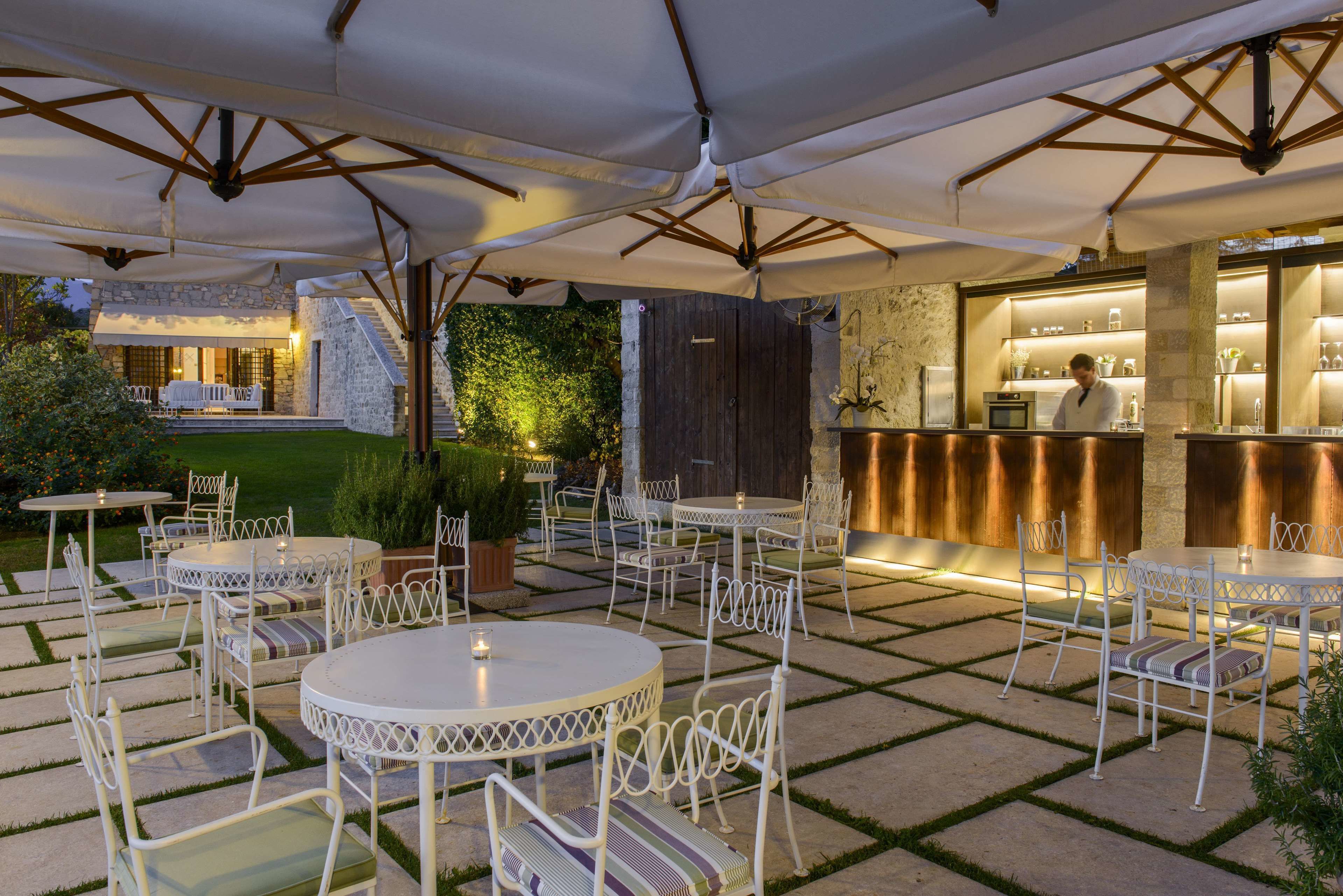 An outdoor patio with round tables and white chairs, a bar area with a counter, and a stone wall with greenery.