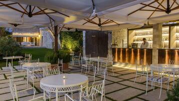 An outdoor patio with round tables and white chairs, a bar area with a counter, and a stone wall with greenery.