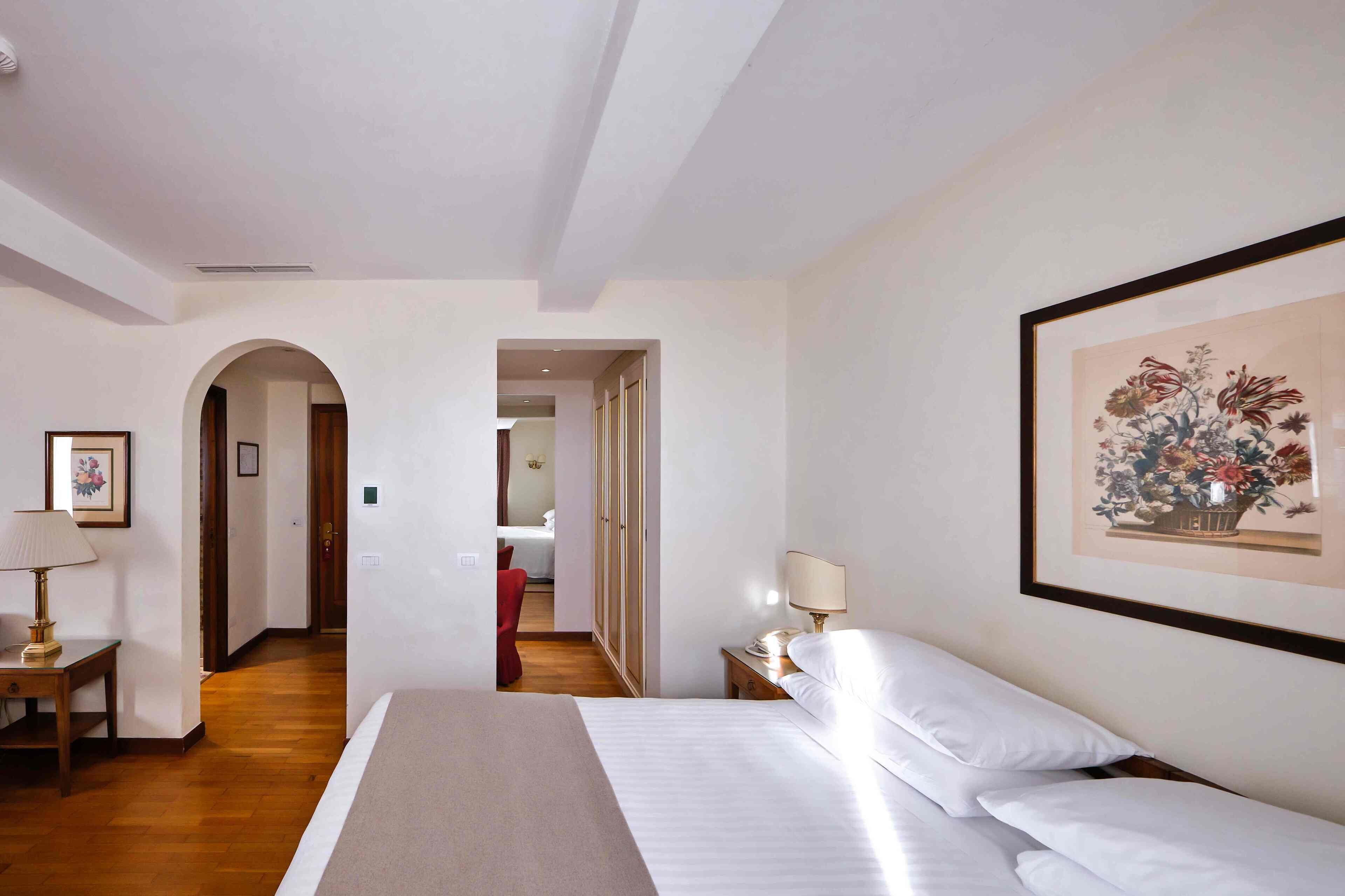 A hotel room with a bed, bedside tables, a lamp, a framed floral artwork, and an arched doorway leading to another room.