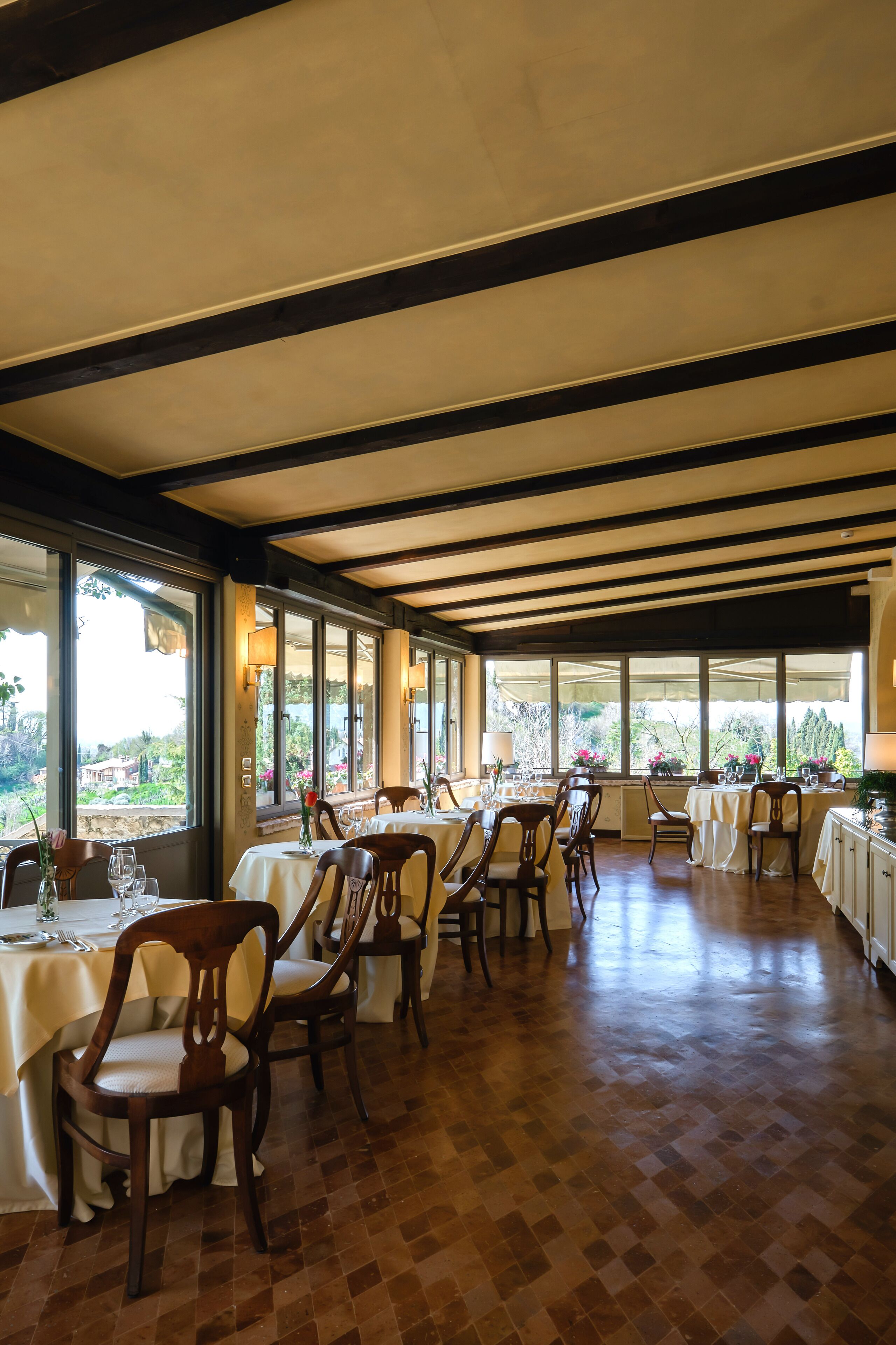 A spacious dining area with multiple tables and chairs, large windows offering a view of the outdoors, and a wooden floor.