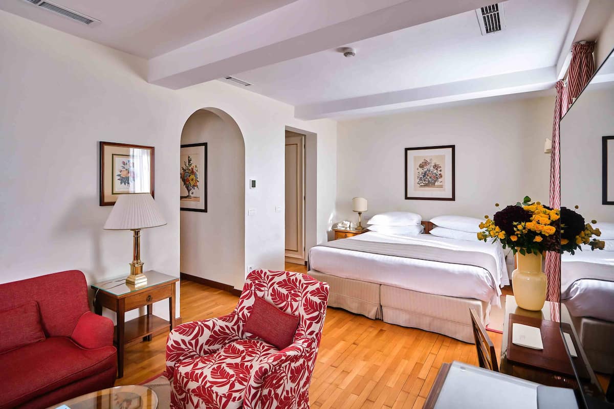 A hotel room with a bed, a red sofa, a patterned armchair, a small table with a lamp, and a vase with flowers.