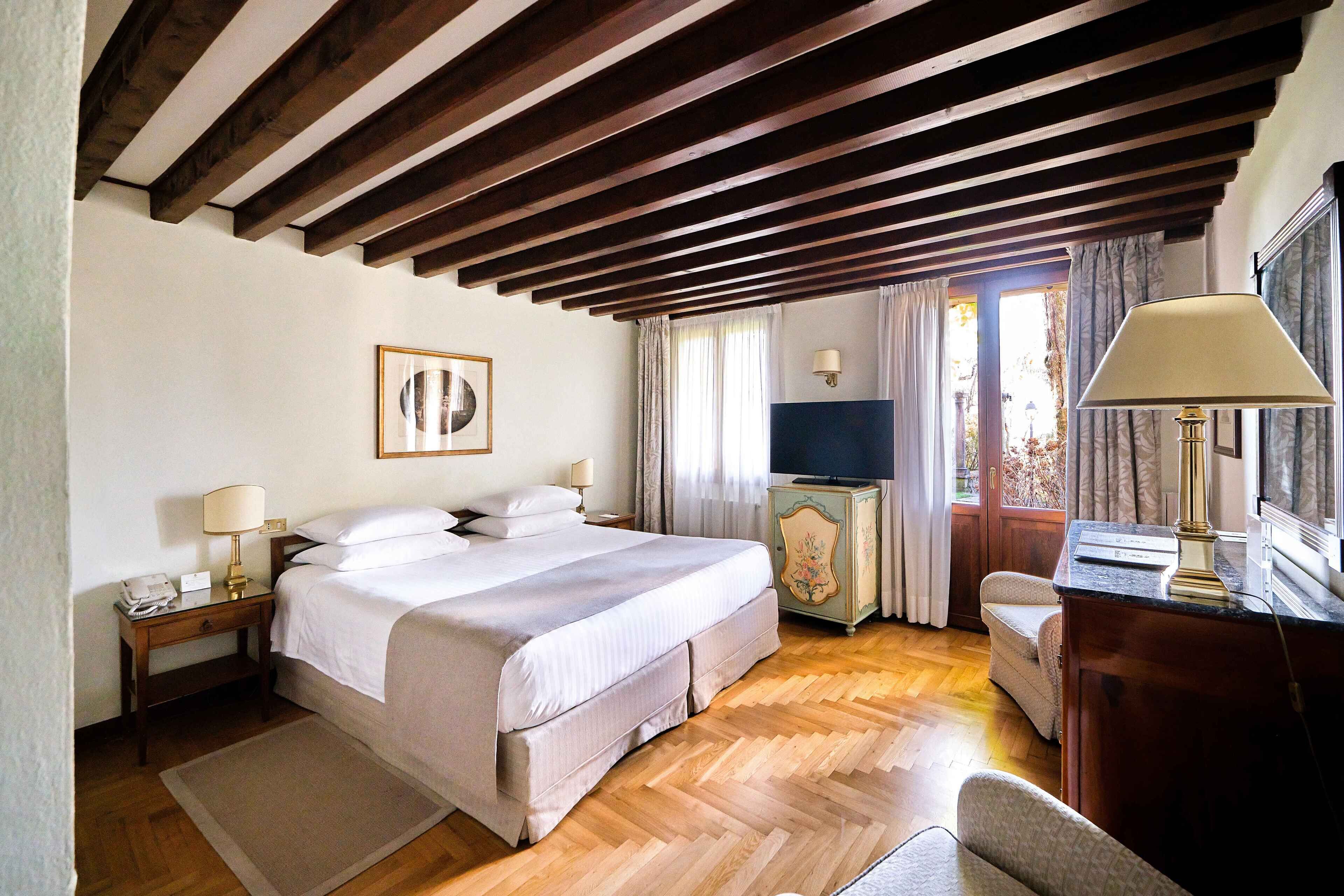Quarto com cama grande, vigas de madeira no teto, escrivaninha com luminária e cómoda.