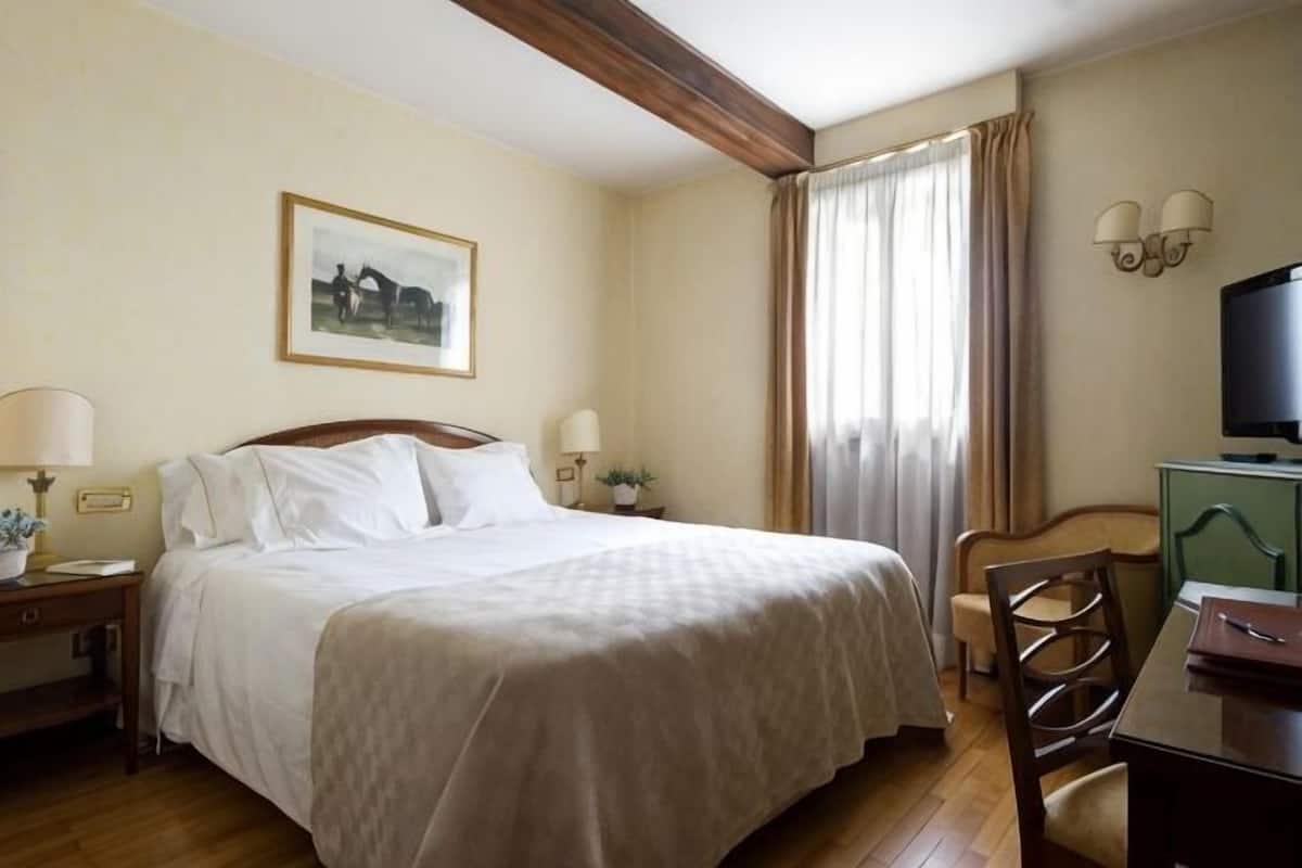 A hotel room with a bed, bedside tables, a desk, a chair, a television, and a window with curtains.