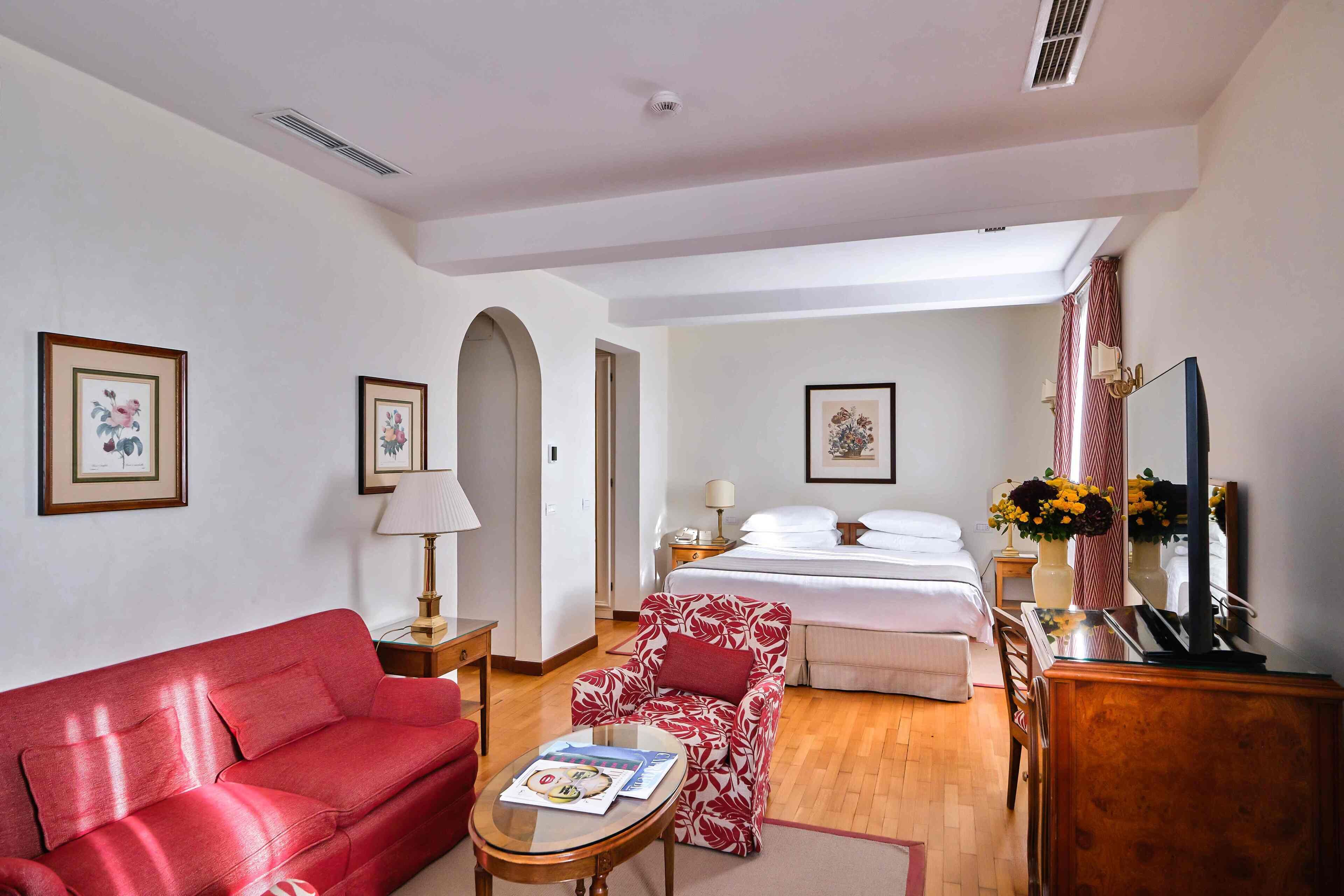 Una habitación de hotel con un sofá rojo, una mesita con una revista, una cama con ropa blanca, un escritorio con una computadora y obras de arte enmarcadas en las paredes.