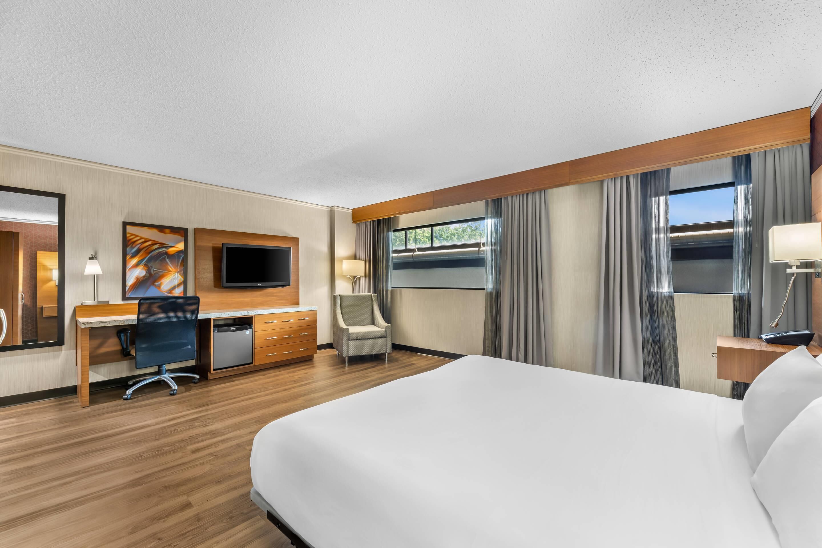 A hotel room with a large bed, a desk with a computer, a chair, a TV, and a window with curtains.