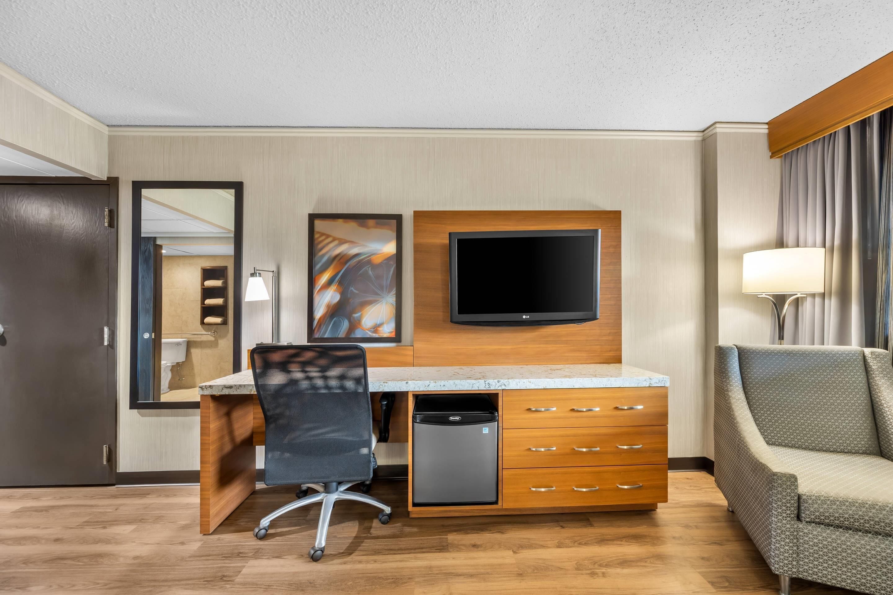 Een hotelkamer met een houten bureau, een zwarte bureaustoel, een televisie, een kleine koelkast, een bank en een spiegel.