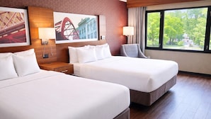 A hotel room with two beds, a nightstand, a lamp, a window with a view of greenery, and a wall with a patterned design and a painting.