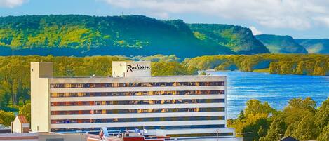 A multi-story hotel with a sign reading 'Radisson,' located near a body of water with a forested hillside in the background.