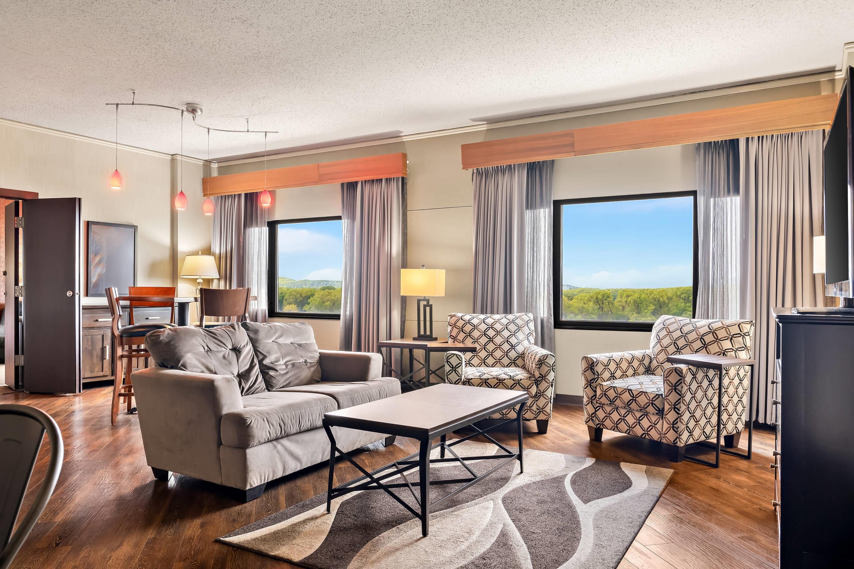 Quarto de hotel com sofá, poltrona, mesa de jantar e vista para o exterior.