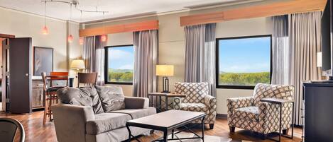 Quarto de hotel com sofá, poltrona, mesa de jantar e vista para o exterior.