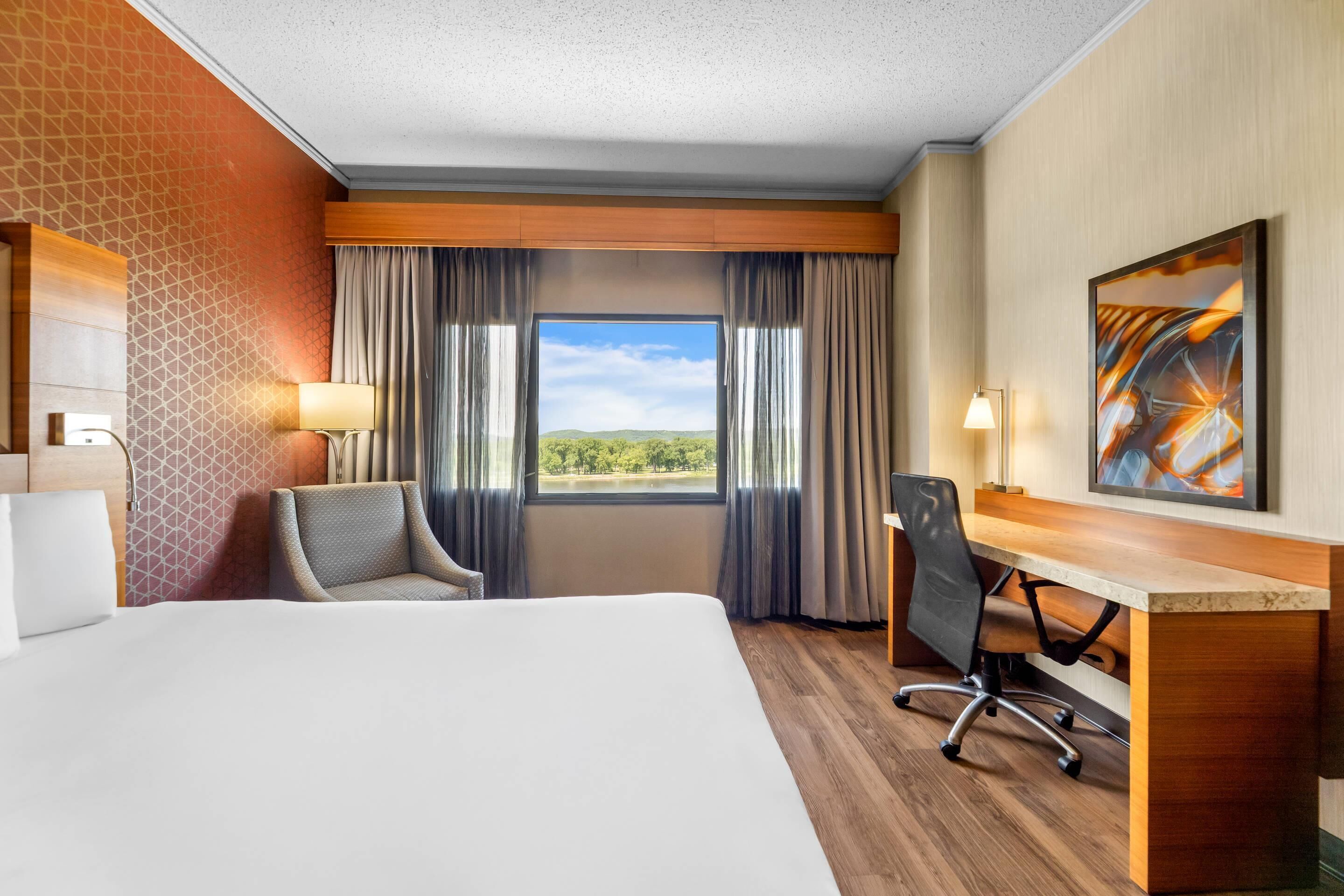 A hotel room with a large bed, a desk, a chair, a window with curtains, and a painting on the wall.