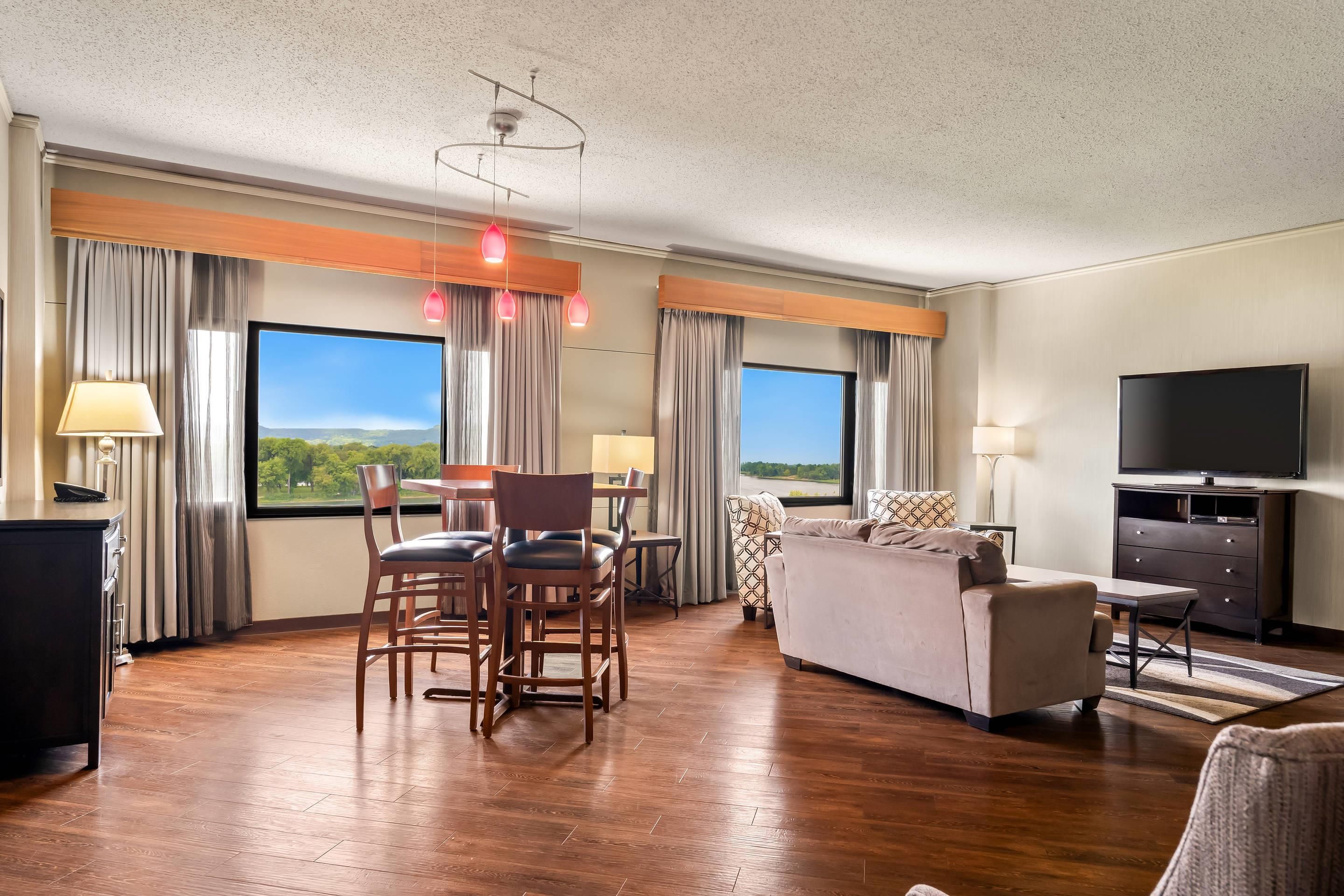 Habitación de hotel con zona de comedor, sofá, televisor y ventanas grandes con cortinas.