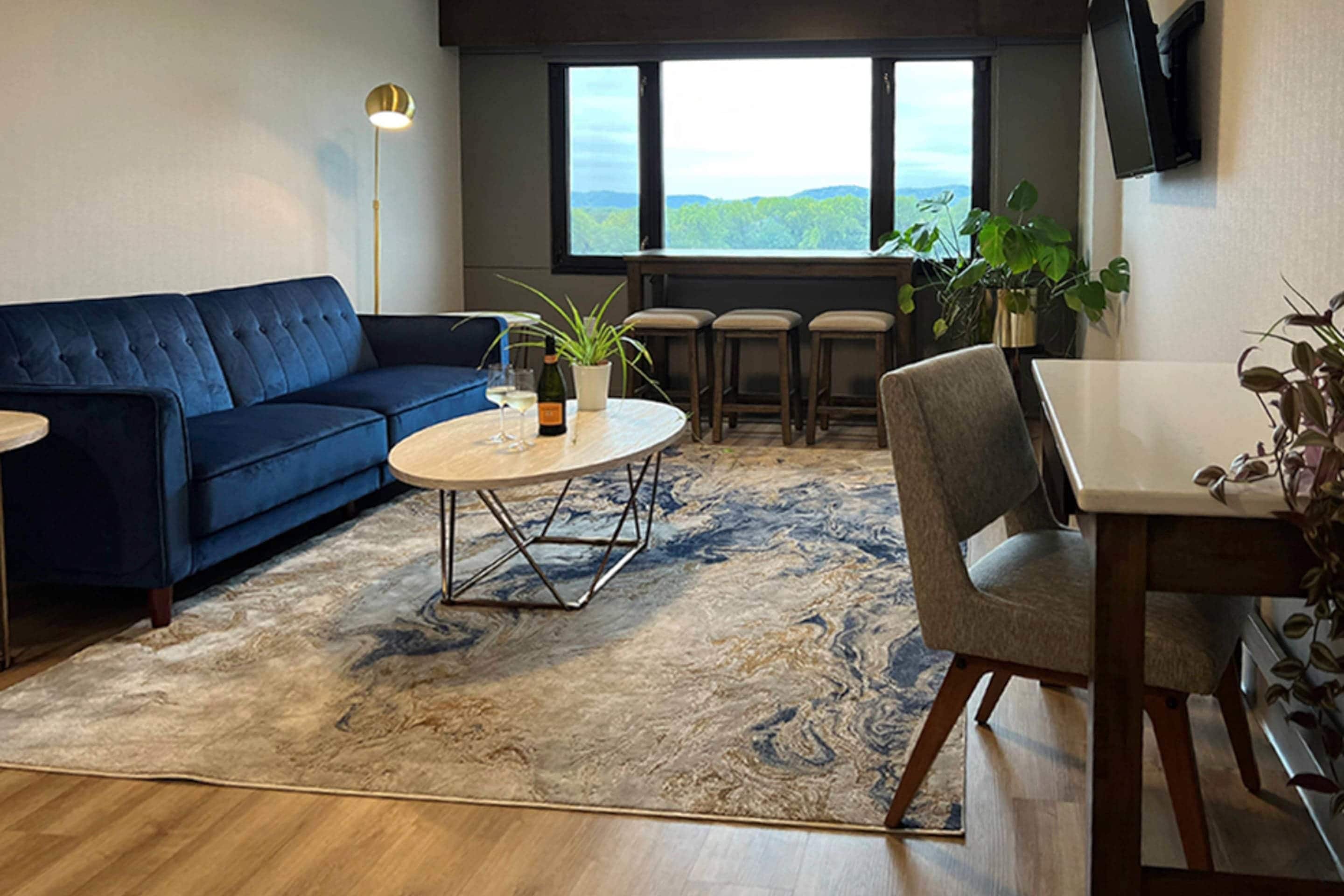 A living room with a blue sofa, a round coffee table, a dining table with chairs, a TV mounted on the wall, and a large window with a view of the outdoors.