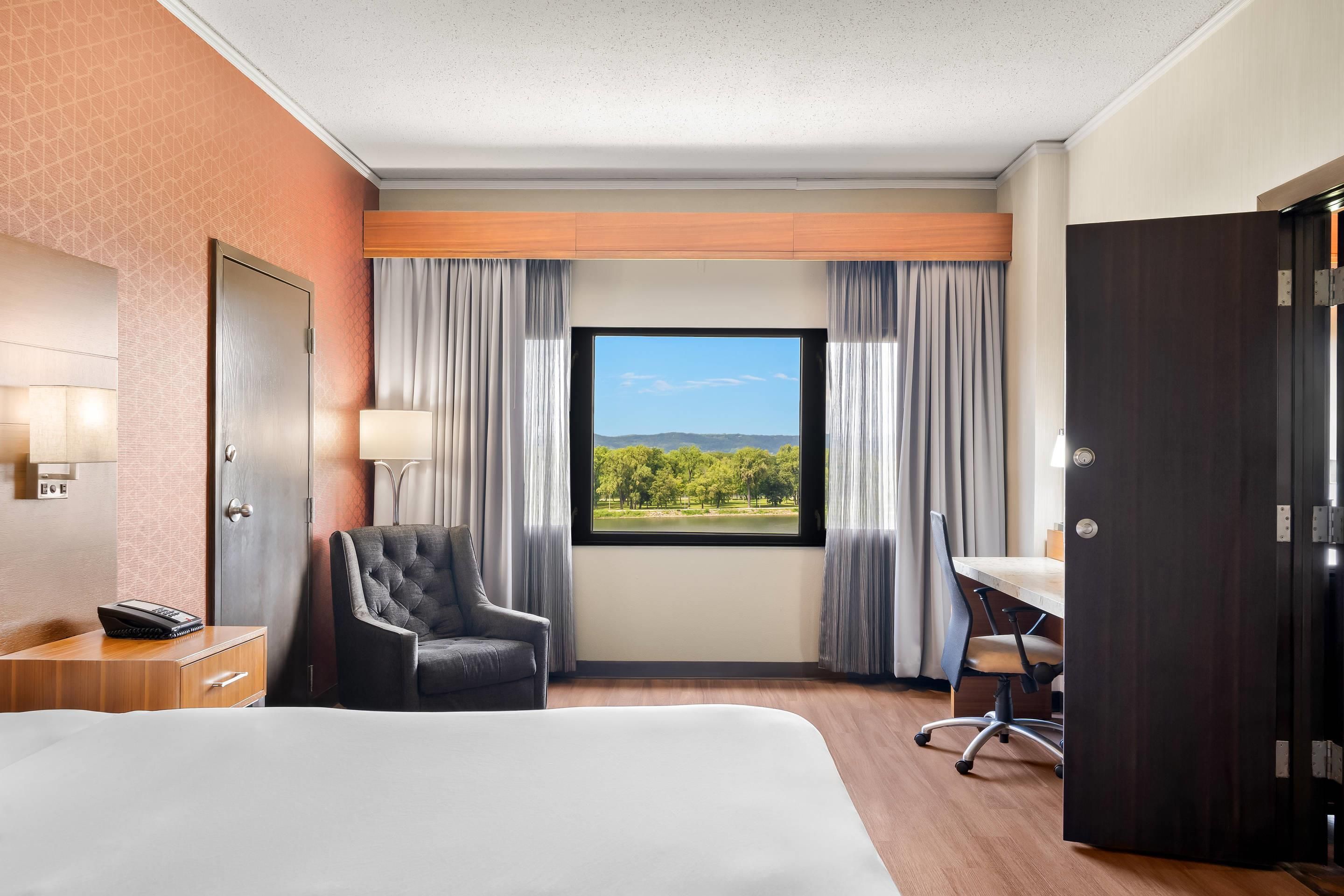 A hotel room with a bed, a desk, a chair, a telephone, and a window with a view of greenery and hills.
