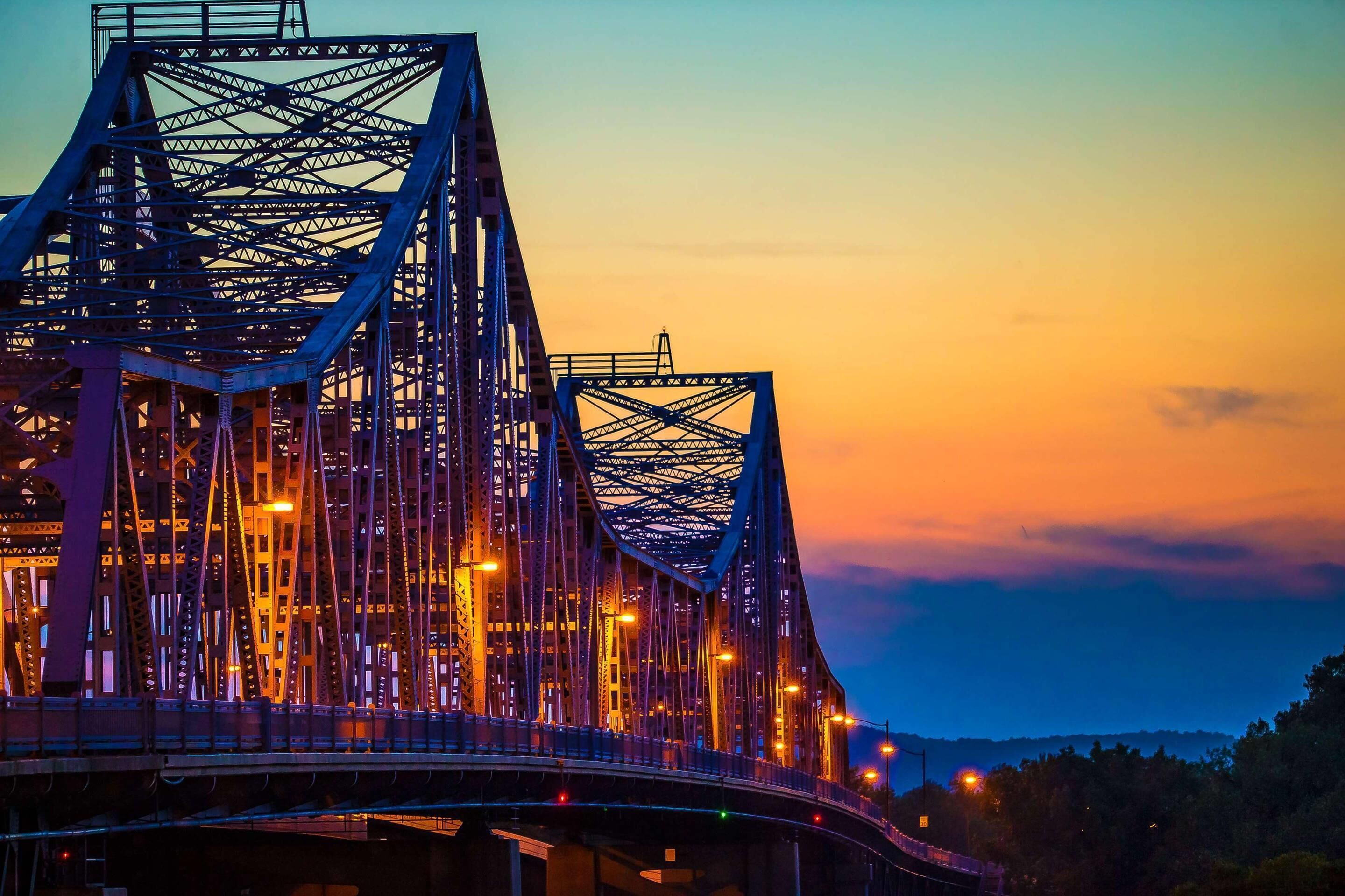A large steel bridge with multiple lanes and a complex truss structure, illuminated by streetlights against a sunset sky.