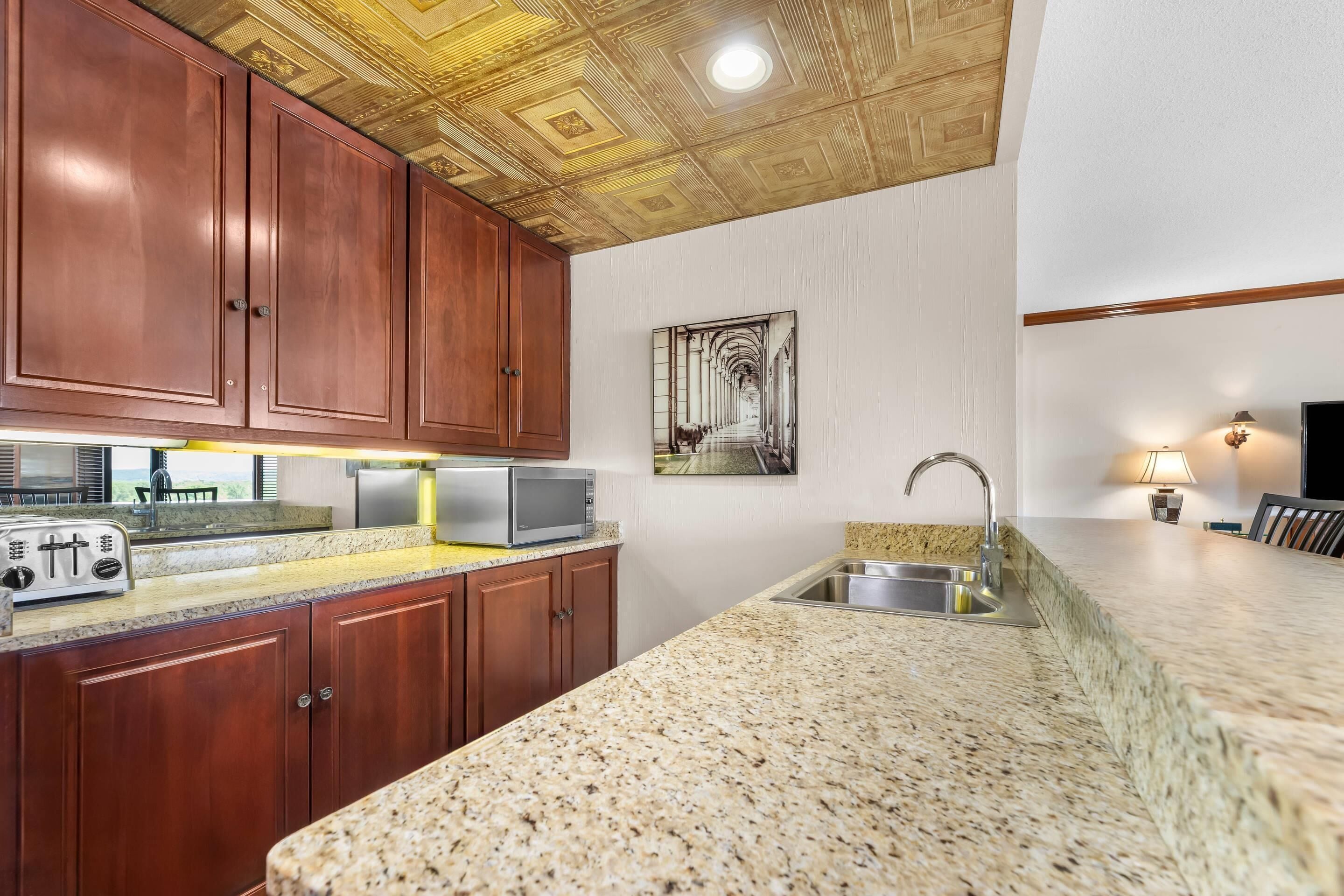 A kitchen with wooden cabinets, a granite countertop, a microwave, a toaster, a sink, and a ceiling with a decorative pattern.