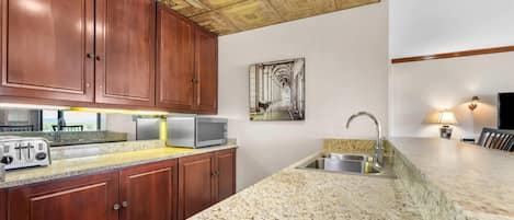 A kitchen with wooden cabinets, a granite countertop, a microwave, a toaster, a sink, and a ceiling with a decorative pattern.