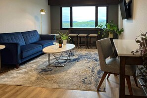 Una sala de estar con un sofá azul, una mesa redonda, una mesa de comedor con sillas, un televisor montado en la pared y un ventanal con vistas al exterior.