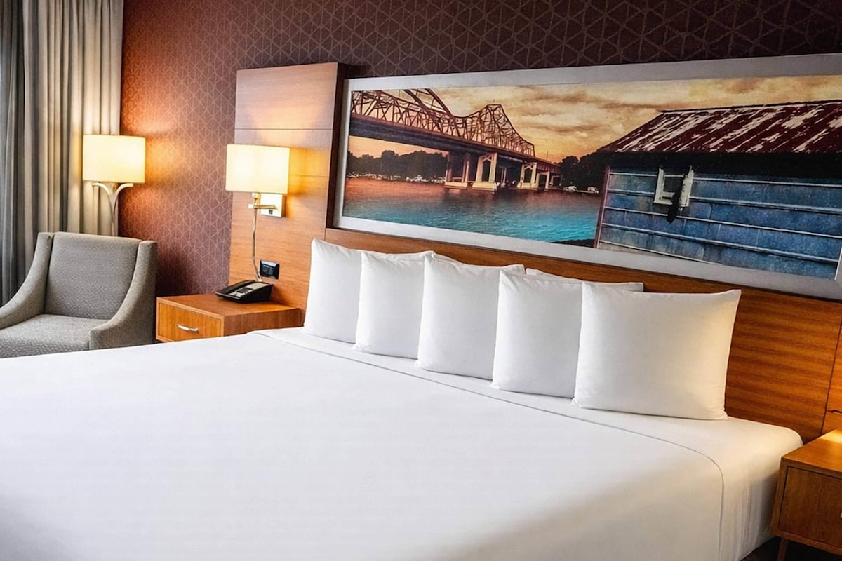 A hotel room with a large bed, a chair, a nightstand, a lamp, and a painting of a bridge.