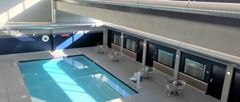 Una piscina interior con un cartel de prohibición de clavados, sillas de descanso y vistas al exterior a través de amplias ventanas.