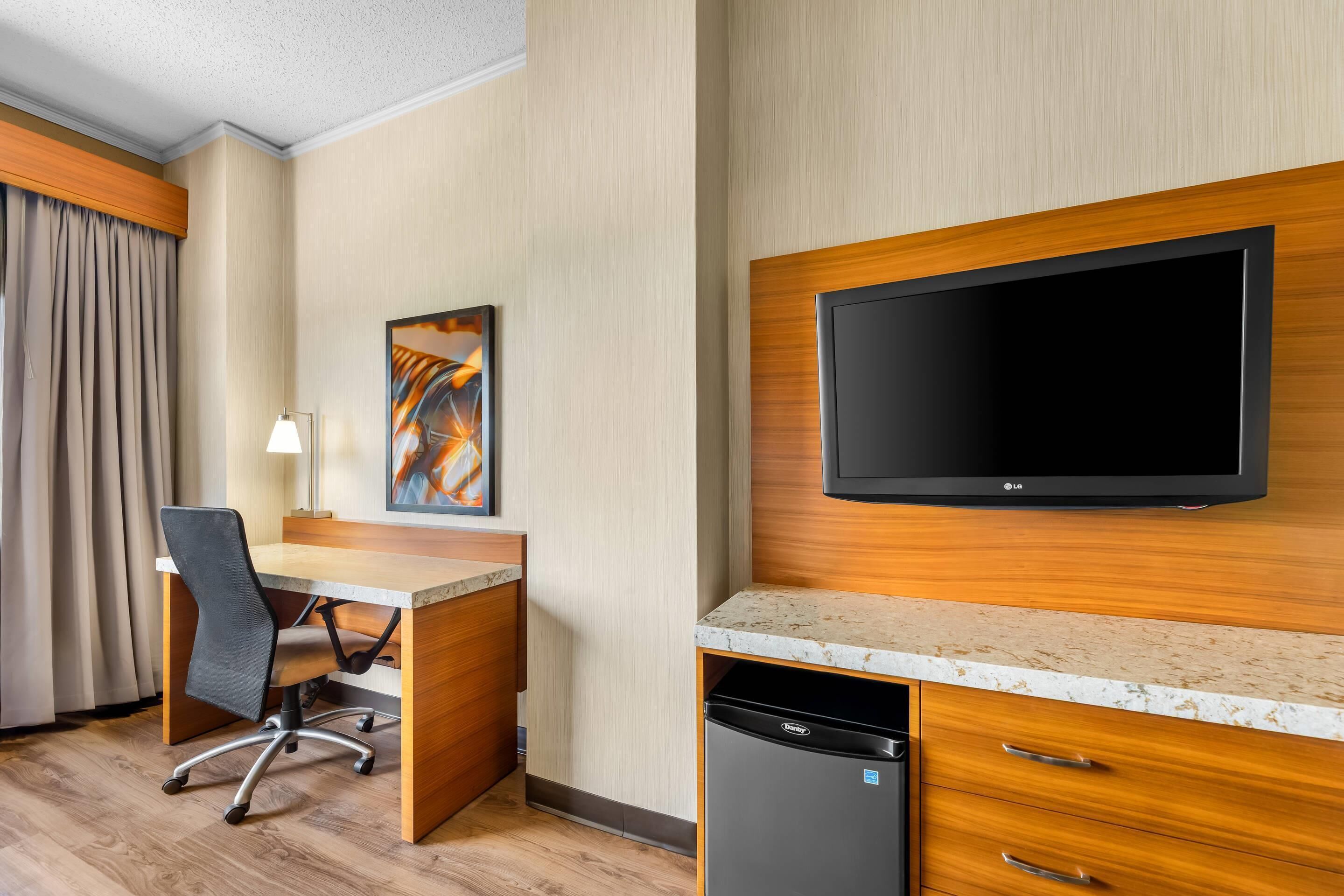 A hotel room with a wooden desk, a chair, a flat-screen TV mounted on the wall, a small black dishwasher, and a painting on the wall.
