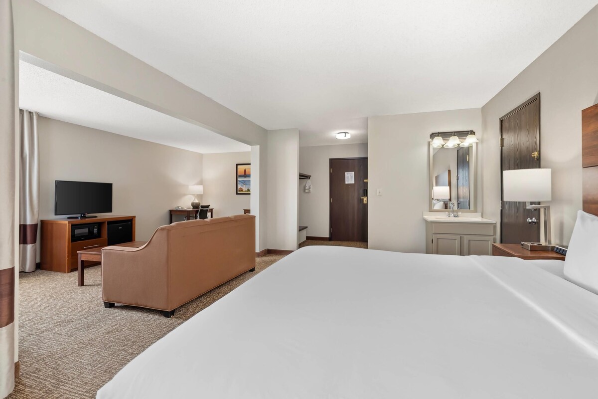 A hotel room with a large bed, a television, a sofa, a desk, and a bathroom with a mirror and sink.