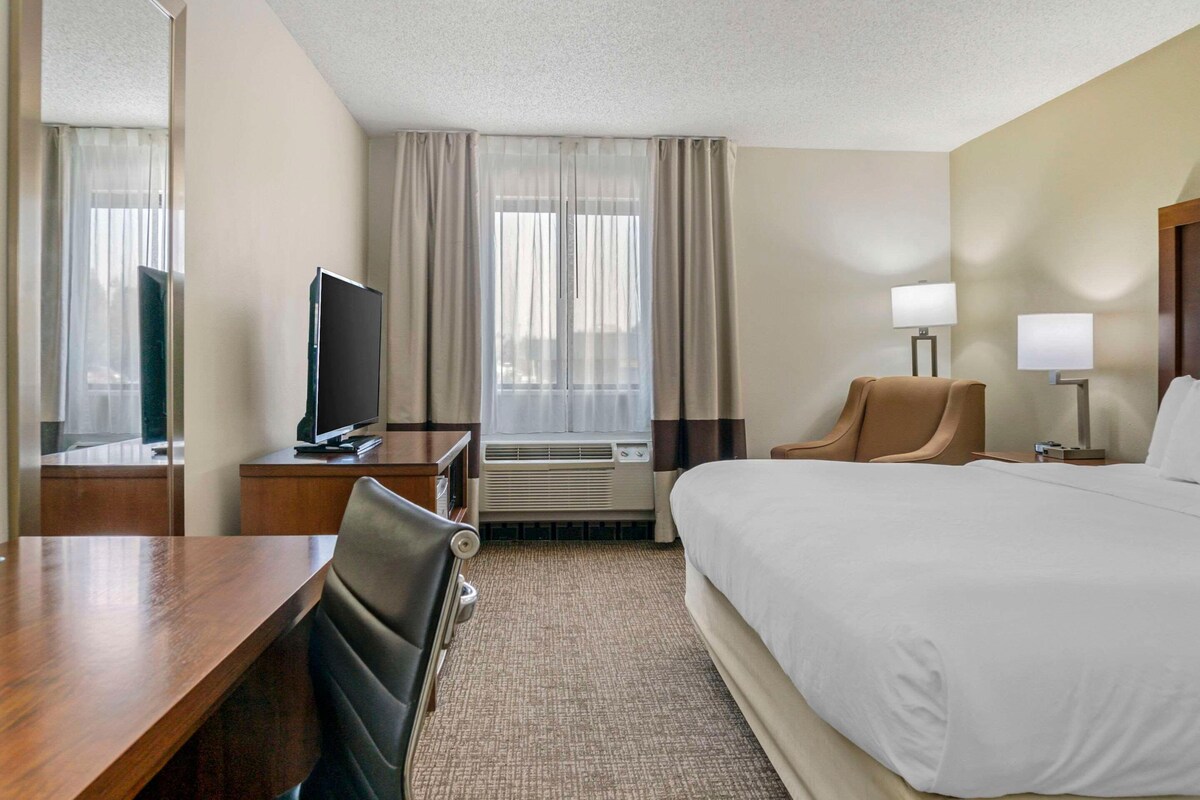A hotel room with a bed, a desk with a television, a chair, a desk lamp, and a window with curtains.