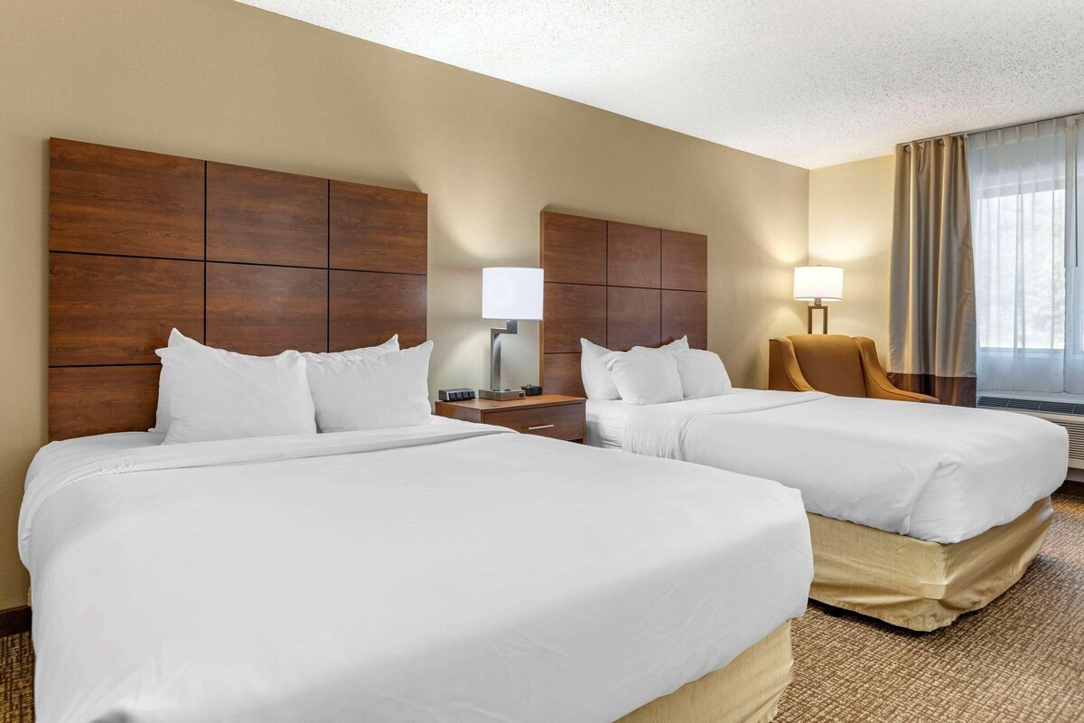 A hotel room with two beds, a nightstand, a lamp, a chair, and a window with curtains.