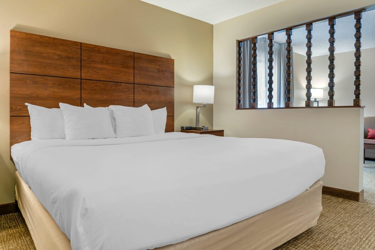 A hotel room with a large bed, a wooden headboard, a nightstand with a lamp, and a mirror on the wall.