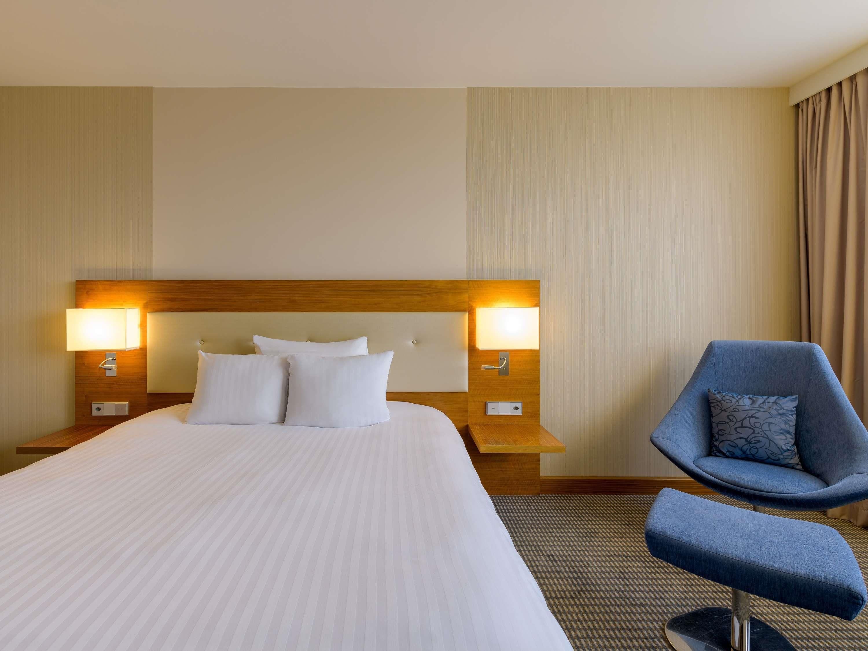 A hotel room with a large bed, two bedside lamps, a blue armchair, and a carpeted floor.