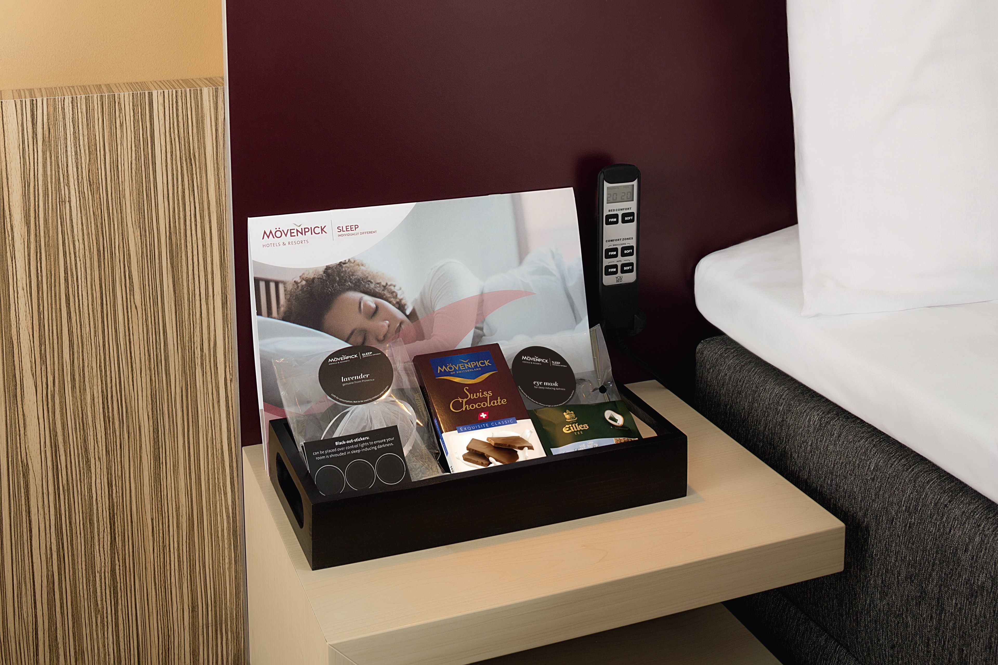 A hotel room with a bedside table holding a sleep brochure, a remote control, and a tray of chocolates.