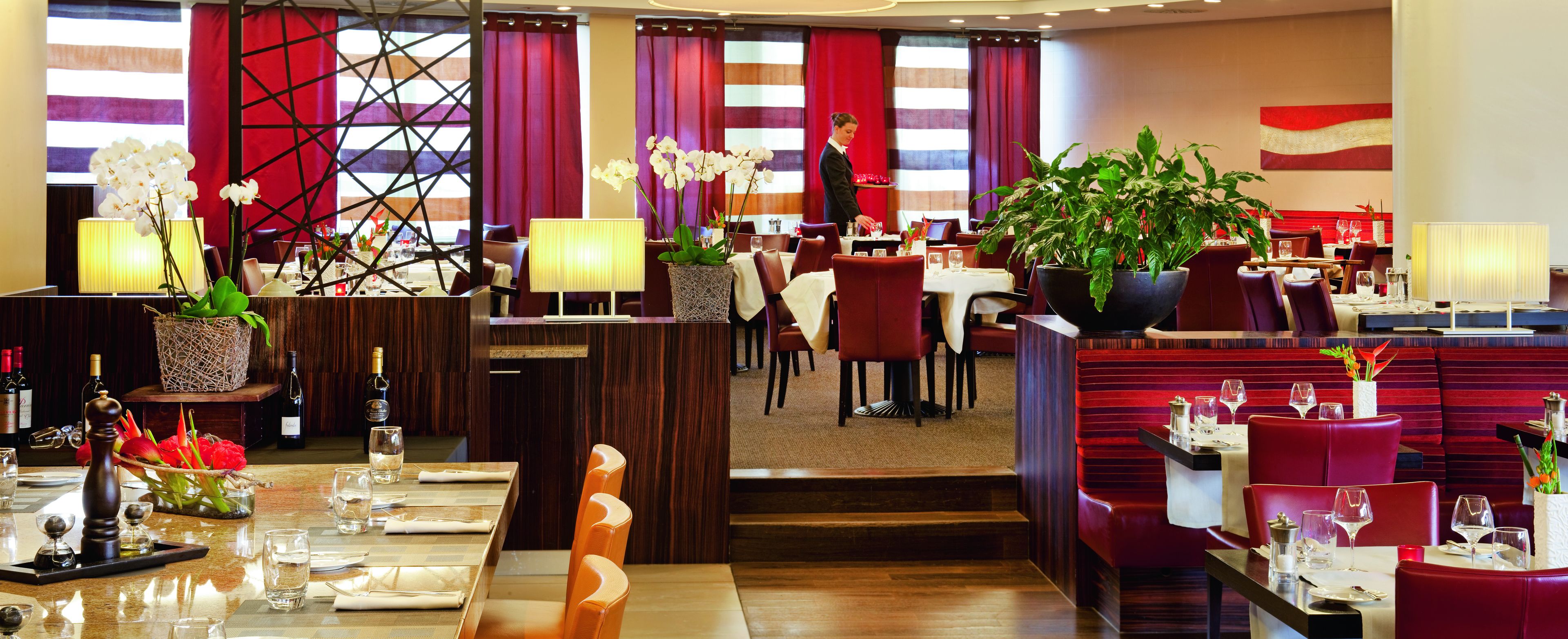 A restaurant with multiple dining tables, red upholstered chairs, and potted plants.