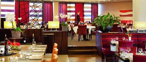 A restaurant with multiple dining tables, red upholstered chairs, and potted plants.