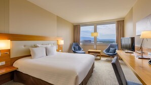 A hotel room with a large bed, two armchairs, a desk with a TV, and a window with a view of the city. - Mövenpick Hotel Geneva (Meyrin)