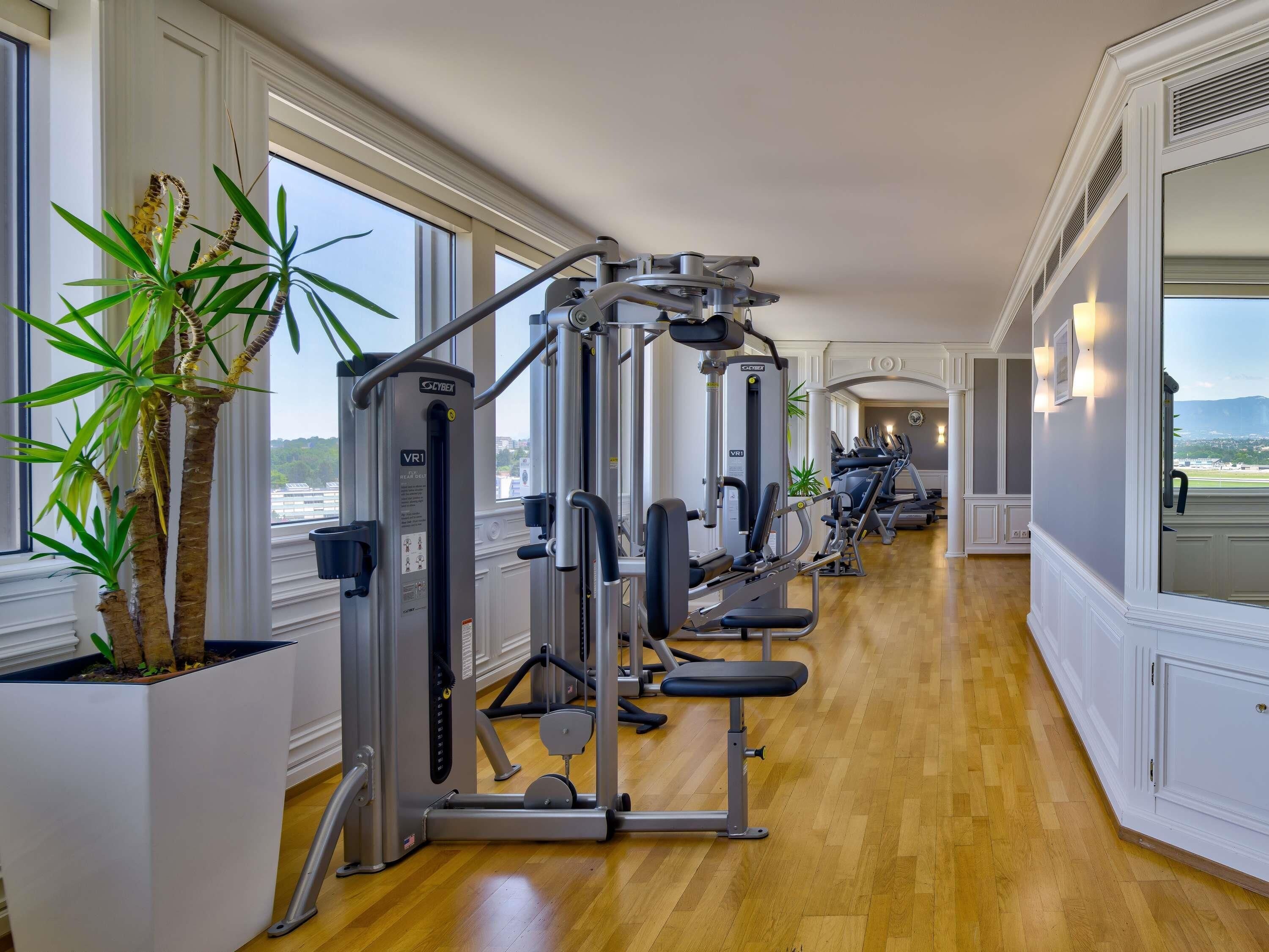 A gym with exercise machines, large windows, and potted plants.