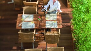 A person reading a magazine at an outdoor wooden table with wicker chairs, surrounded by greenery. - Mövenpick Hotel Geneva (Meyrin)