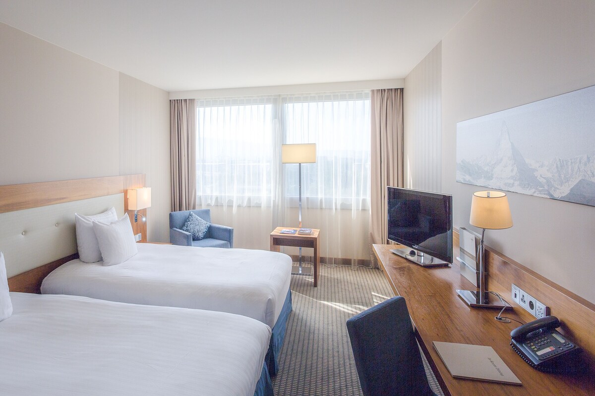 A hotel room with two beds, a desk with a TV, a chair, and a lamp.