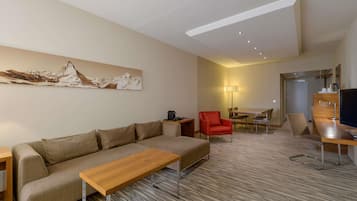 Quarto de hotel moderno com sofá, mesa de centro de madeira, poltrona vermelha, mesa de jantar com cadeiras e televisão em suporte de madeira.