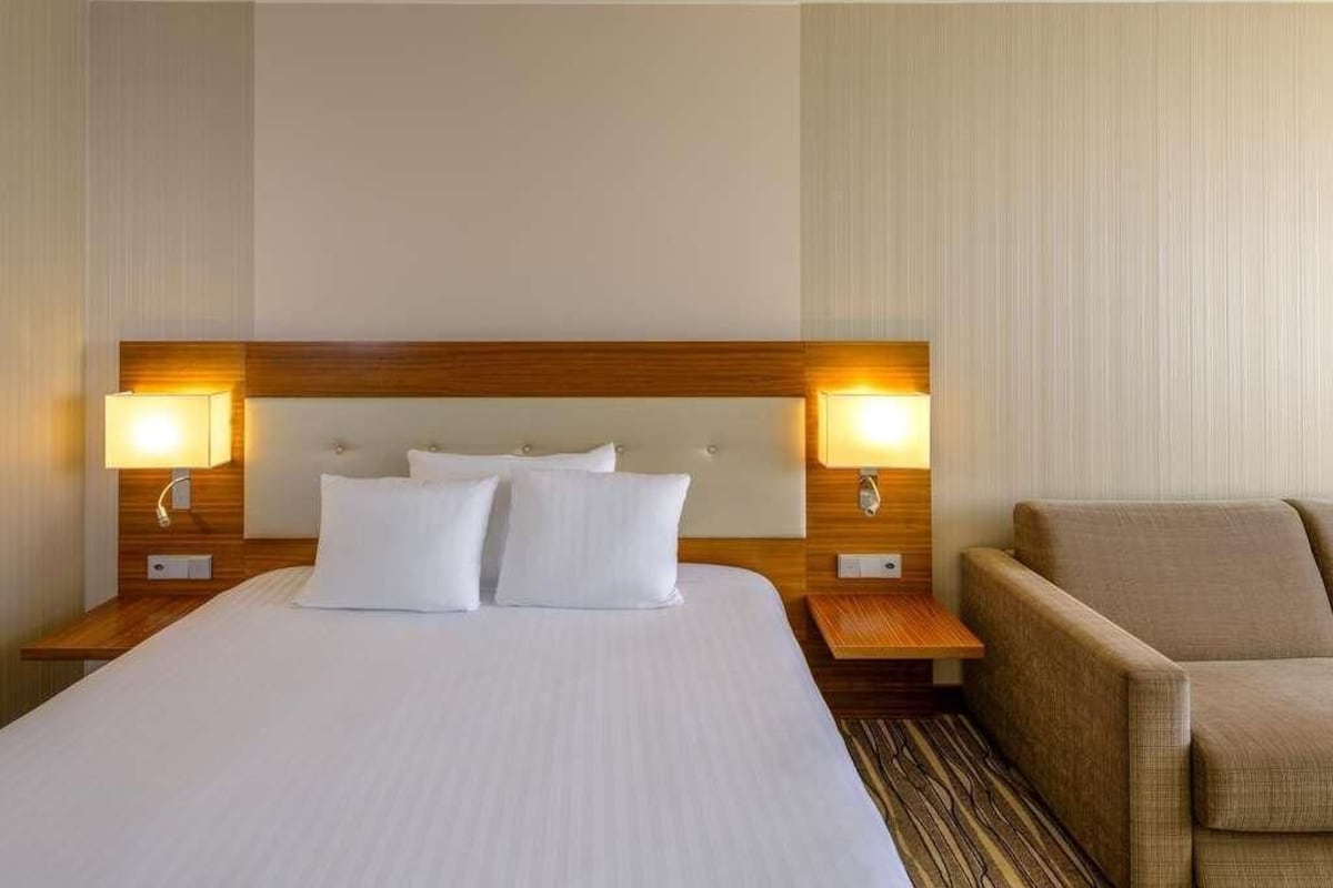 A hotel room with a large bed, two bedside lamps, a wooden headboard, a brown armchair, and a patterned carpet.